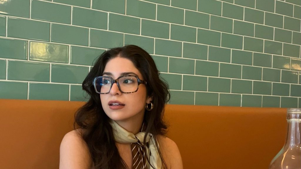 woman sitting at a booth wearing oversized glasses and a scarf tied around her neck